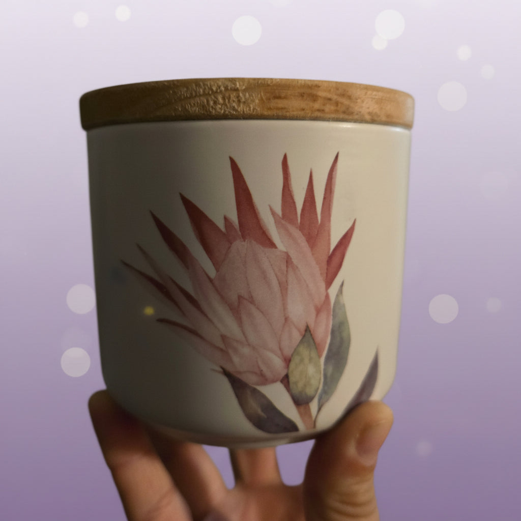 A hand holding a white ceramic jar with a natural bamboo lid and a painted pink flower, set against a blurred indoor background.
