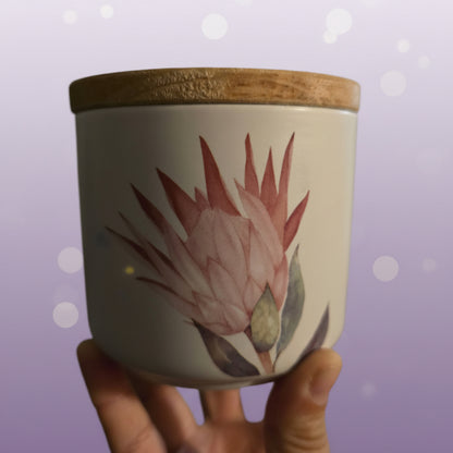 A hand holding a white ceramic jar with a natural bamboo lid and a painted pink flower, set against a blurred indoor background.