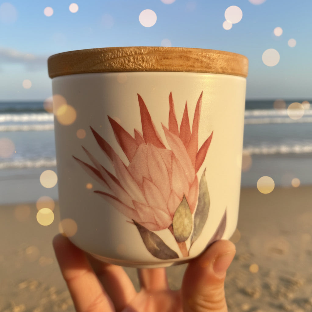 A hand holding a white ceramic jar with a light brown wooden lid and a pink and green protea flower illustration on the front, set against a soft beach vibe