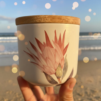 A hand holding a white ceramic jar with a light brown wooden lid and a pink and green protea flower illustration on the front, set against a soft beach vibe